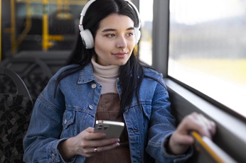 A dark haired woman sitting on a train, looking out of the window, wearing headphones and holding a phone