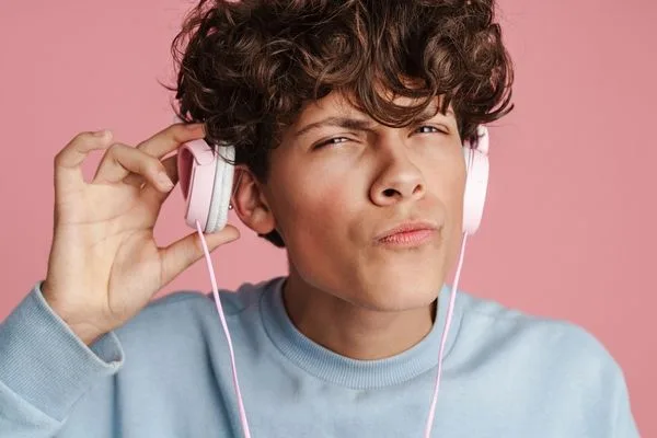 Photo of a white man wearing headphones, lifting one side away from his ear with a confused face