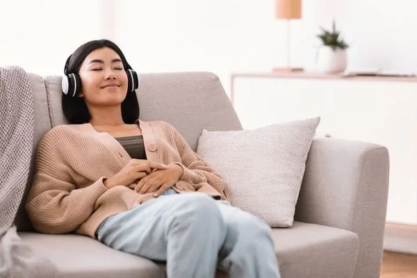 An Asian woman resting on a sofa, eyes closed, headphones on