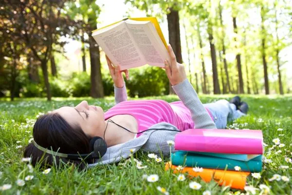 A white woman with long dark hair lying in the grass and reading, a stack of colorful books next to her and headphones on her ears