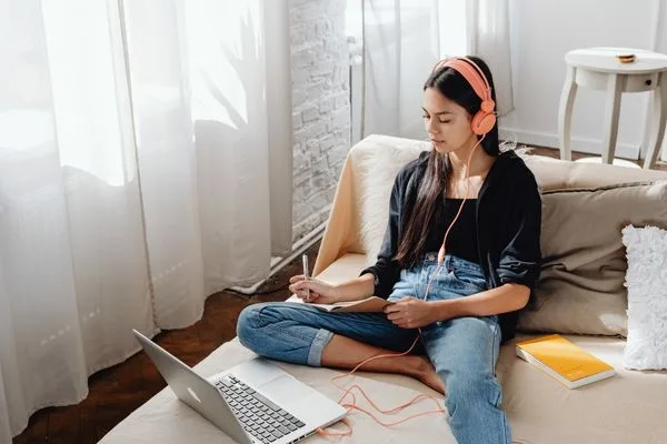 Photo of a white woman with long dark hair sitting on a sofa with a notebook and pen in hand, headphones on her ears