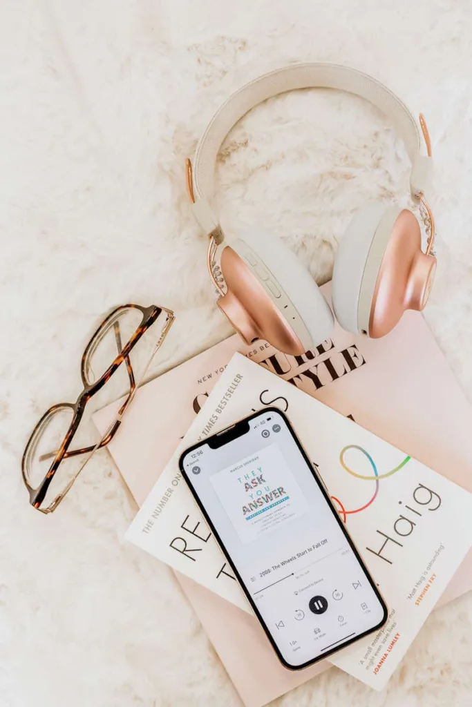 Starting with audiobooks: Flatlay of reading glasses, a paperback book written by Matt Haig, a phone playing an audiobook and white and rose gold headphones