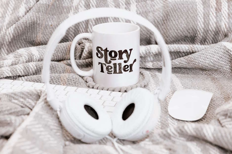 How to listen to audiobooks: Flatlay showing white headphones and a keyboard lying on a grey blanket, and a mug with the words "story teller"