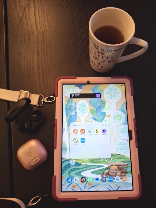 About Me 2 A big cup of tea, a tablet showing audiobook apps, and a pink and a black case of earbuds